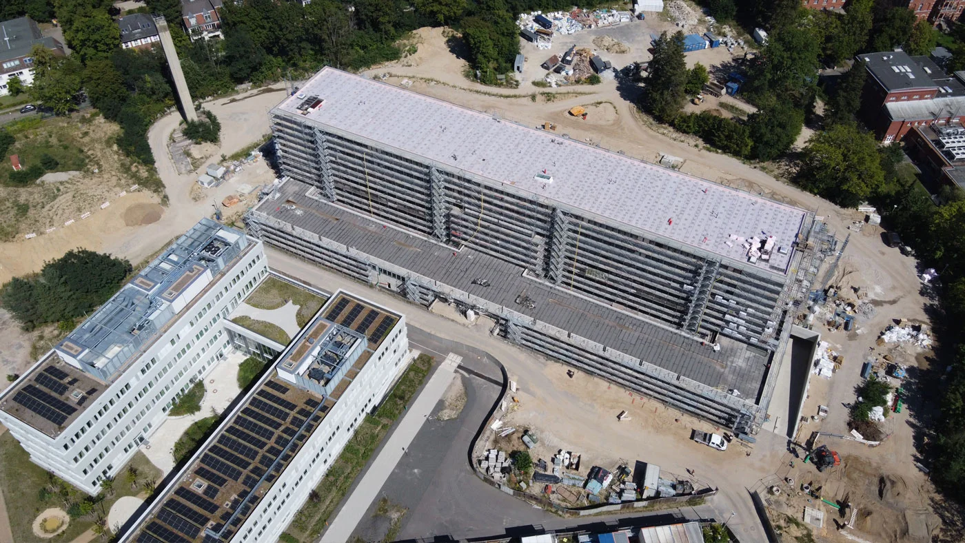 Luftaufnahme einer Baustelle mit einem langgestreckten, mehrstöckigen Gebäude und einem angrenzenden, weißen Bürogebäude mit Solarpaneelen auf dem Dach